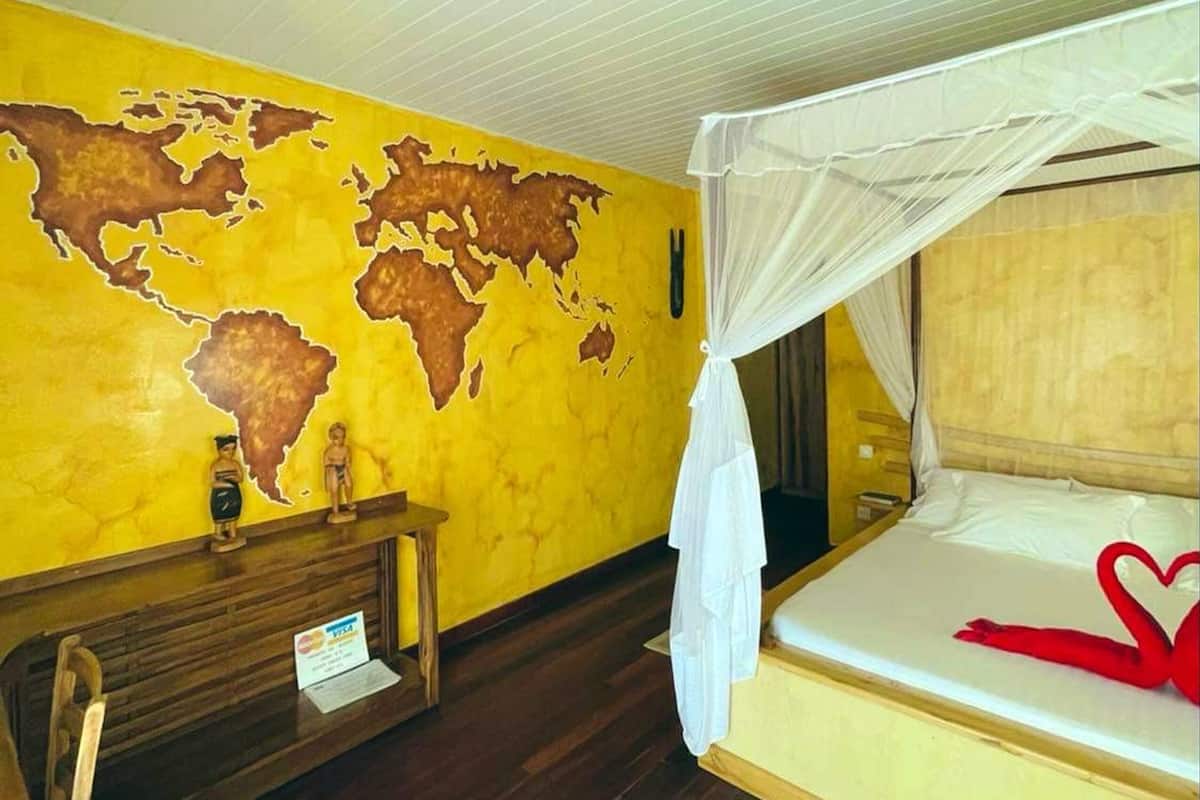 Quarto de hotel com uma cama, uma escrivaninha, uma cadeira, um mapa na parede e mosquiteiro sobre a cama.