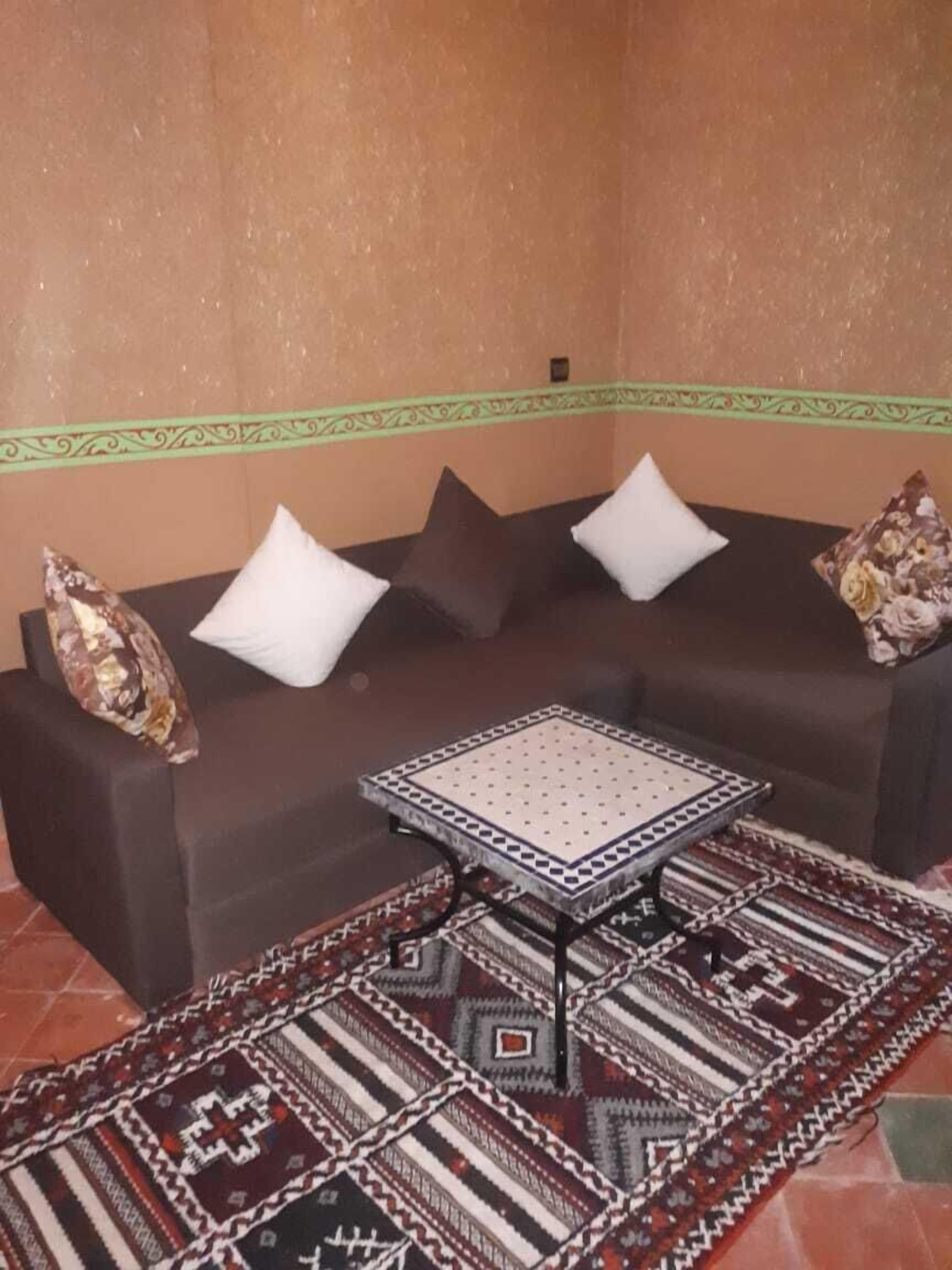 Una habitación de esquina con un sofá marrón oscuro, cojines blancos y marrones con estampado, una mesita cuadrada con superficie estampada y una alfombra decorativa con diseños geométricos y tribales.