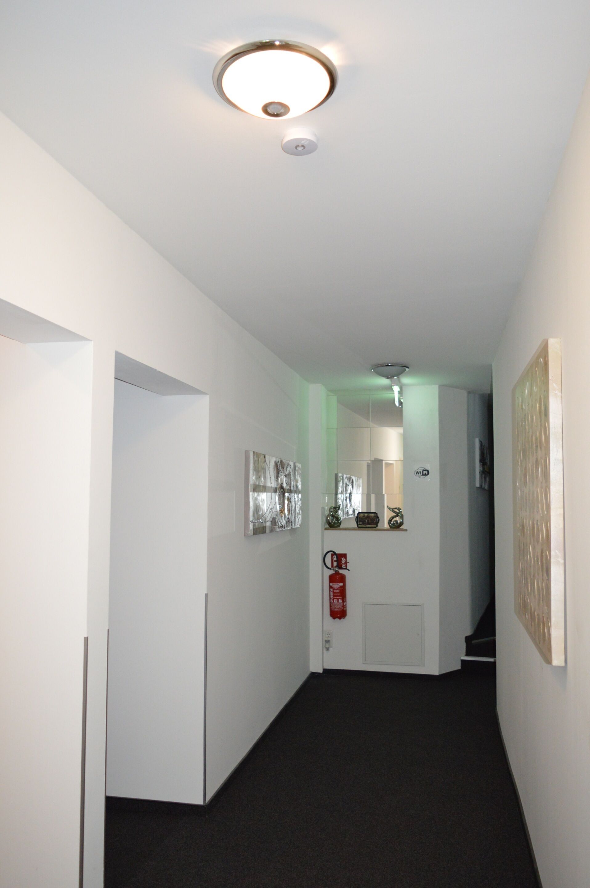 A hallway with white walls, a black carpet, a fire extinguisher, and a door leading to a staircase.