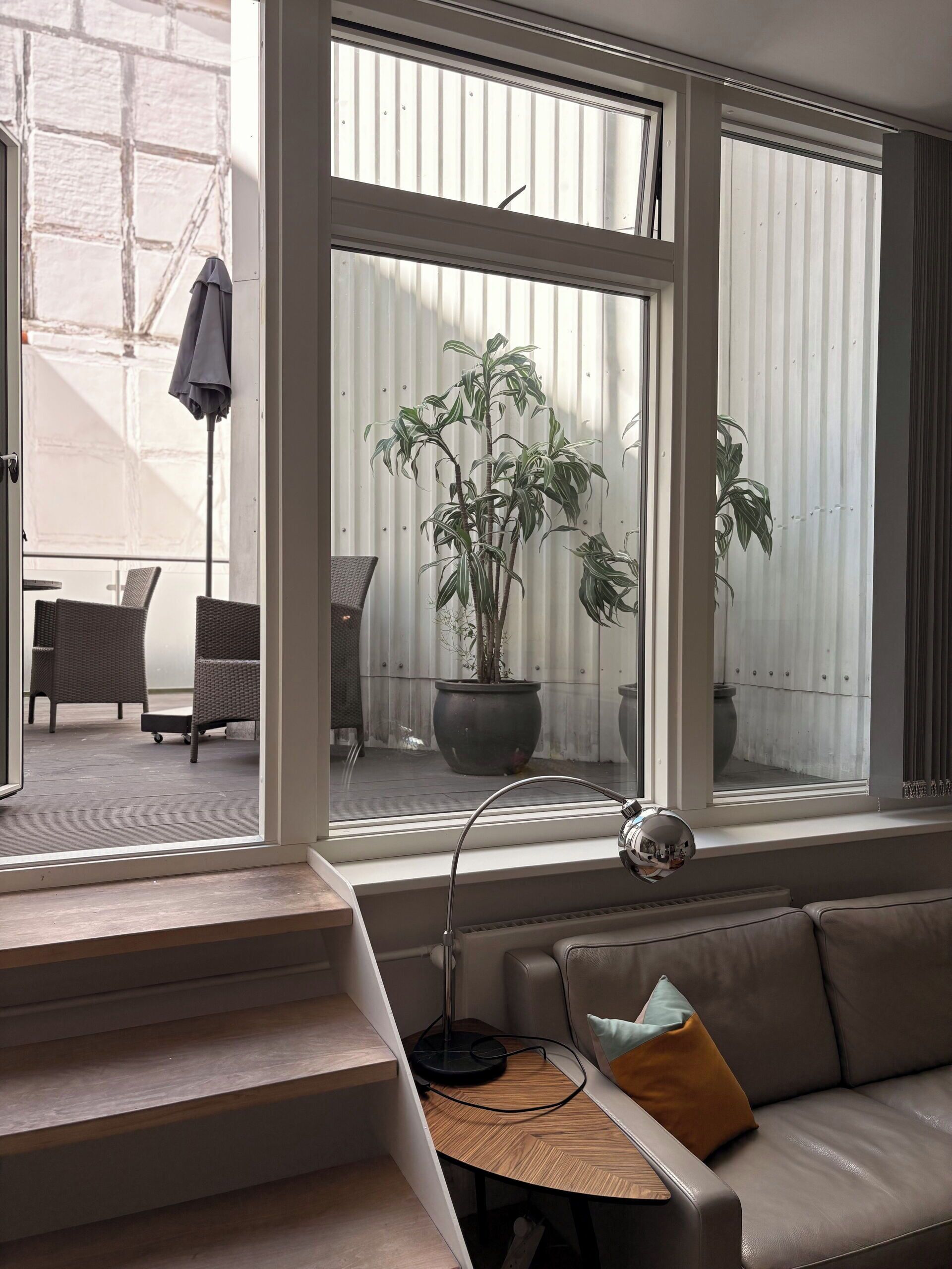 A modern living room with a sofa, a coffee table, and a large window overlooking a balcony with outdoor furniture.