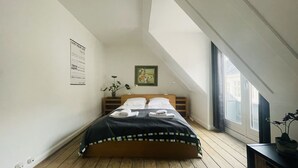 Een slaapkamer met een schuine plafond, een bed met netjes opgevouwen handdoeken, een ingelijst foto aan de muur, en een raam met gordijnen.