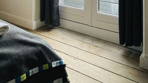 Een kamer met een houten vloer, een raam met donkere gordijnen en uitzicht op een gebouw met een witte gevel en een pannendak.