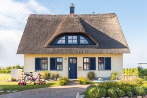 Une maison avec un toit de chaume, une porte bleue et un petit jardin où se trouvent un vélo et une voiture jouet.