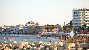 Une plage où l’on voit des gens nager et prendre des bains de soleil, un mur de pierre et des bâtiments en arrière-plan.