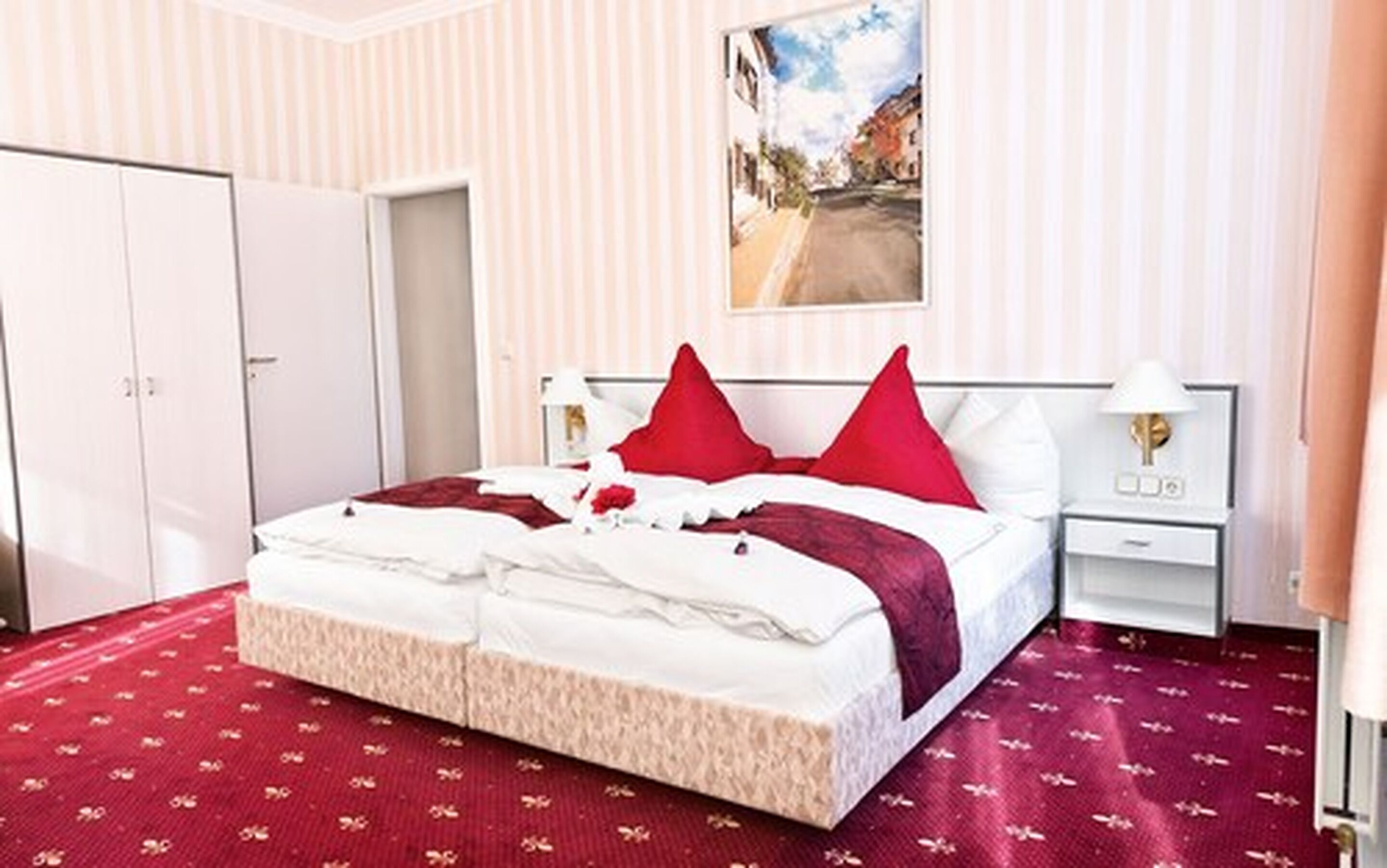A hotel room with a large bed, two red pillows, a white headboard, a nightstand with a lamp, and a painting of a street scene on the wall.