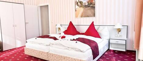 Habitación de hotel con una cama grande, dos almohadas rojas, un cabecero blanco, una mesita de noche con una lámpara y un cuadro de una escena callejera en la pared.