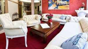 A hotel lobby with a red carpet, white upholstered sofas, a wooden coffee table, and a painting of a coastal scene. - Parkhotel Meerane (Meerane)
