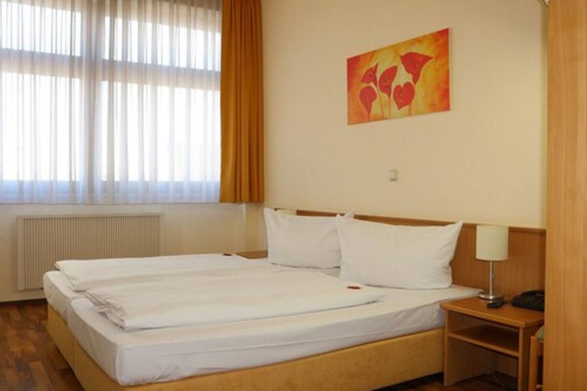 A hotel room with two single beds, a bedside table with a lamp, a radiator, and a window with curtains.