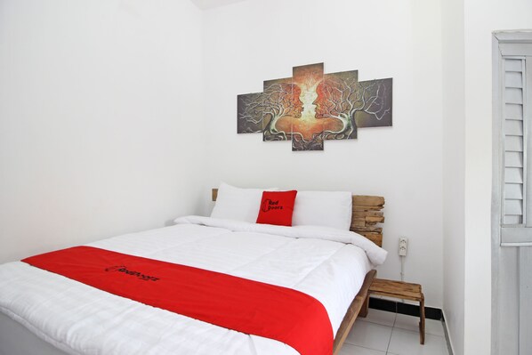 Habitación de hotel con cama, colcha roja y blanca, cabecera de madera, mesita de noche pequeña de madera y una obra de arte colgada en la pared.