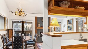 A dining area with a round table and chairs, a chandelier, and a view through the window. - Outpost: Hollyhock 2025 (Wilson)