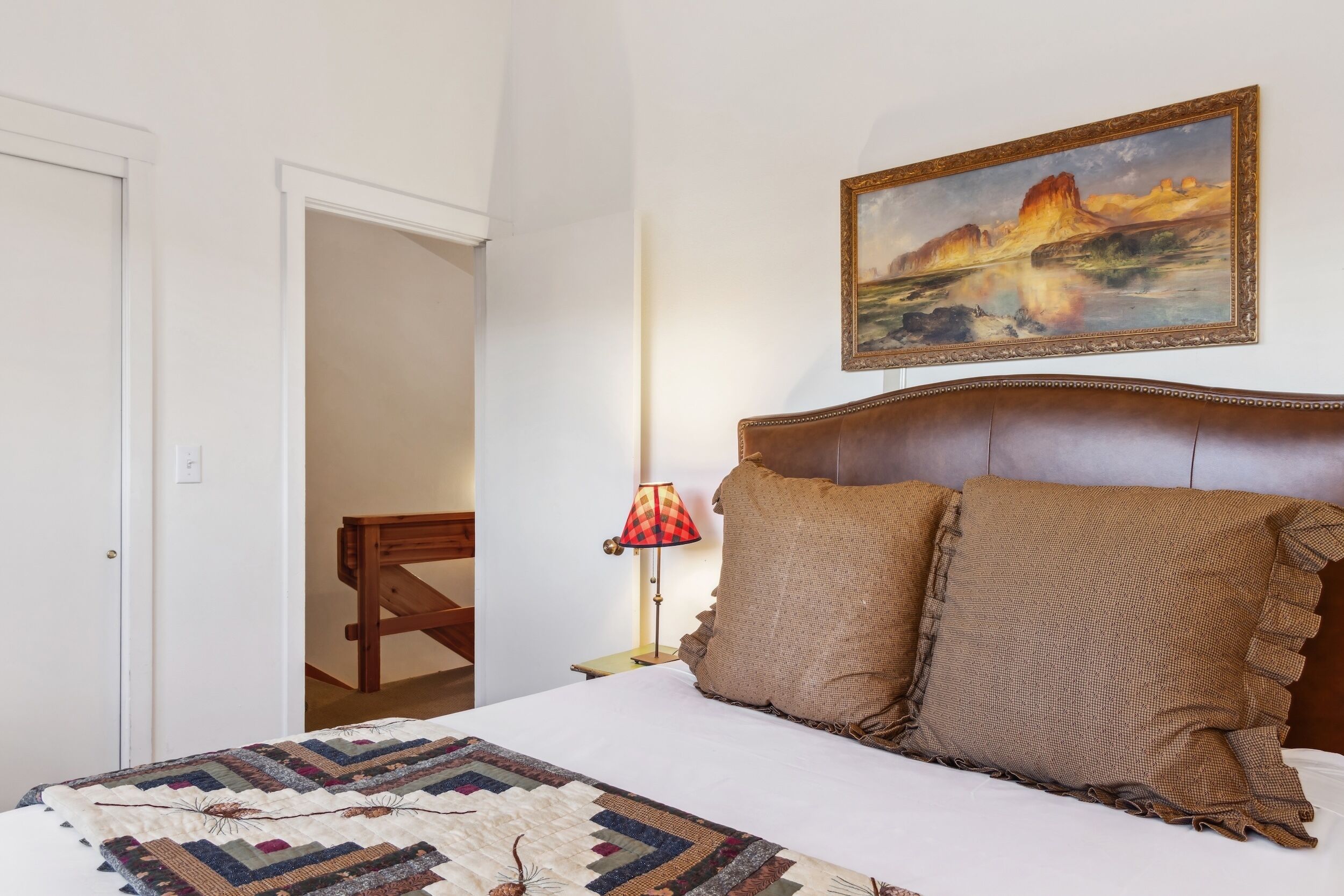 A bedroom with a large bed, a painting on the wall, a lamp, and a wooden chair.