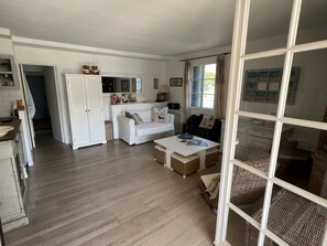 Ett rymligt vardagsrum med en vit soffa, ett soffbord och ett matbord. Här finns också ett köksområde med en diskho och skåp.