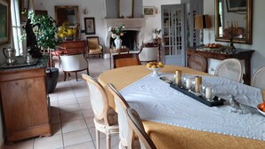 Un'ampia sala da pranzo con un grande tavolo apparecchiato per sei, circondato da sedie in legno. Si nota un caminetto sullo sfondo, e la stanza presenta uno stile architettonico classico con travi a vista e pavimento piastrellato.
