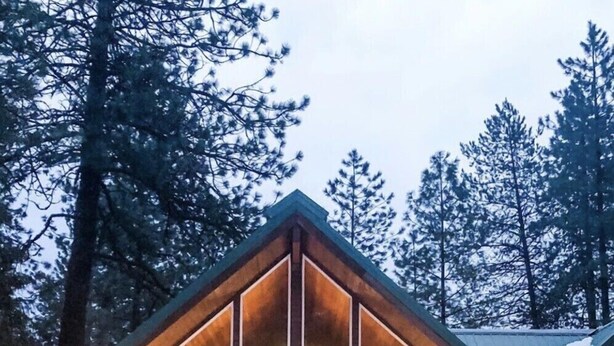 A wooden cabin with a triangular roof and large windows, surrounded by snow and trees.