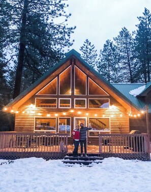 Een houten hut met een puntig dak en grote ramen, omgeven door sneeuw en bomen.