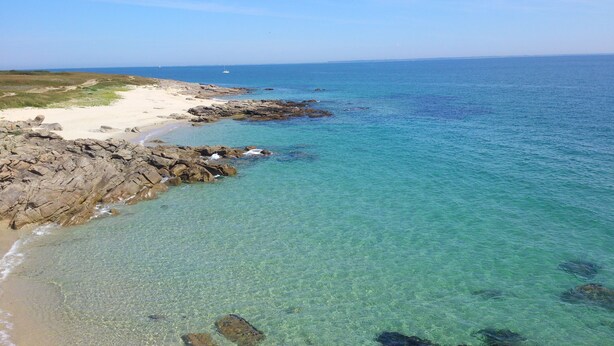 Una playa costera con aguas cristalinas, costa rocosa y un velero a lo lejos.