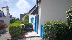 Un sendero estrecho que lleva a un edificio con contraventanas azules y fachada blanca. Hay setos bien cuidados y plantas con flores a lo largo del sendero.