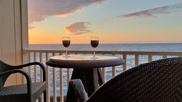 A balcony with wicker chairs and a small round table, two glasses of red wine, and a view of the ocean at sunset. - Amazing Direct Ocean Front Condo - Beautiful Sunsets, watch dolphins and whales (Kailua-Kona)