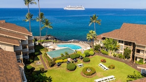 A resort with a swimming pool, palm trees, and a view of the ocean and a cruise ship. - Amazing Direct Ocean Front Condo - Beautiful Sunsets, watch dolphins and whales (Kailua-Kona)