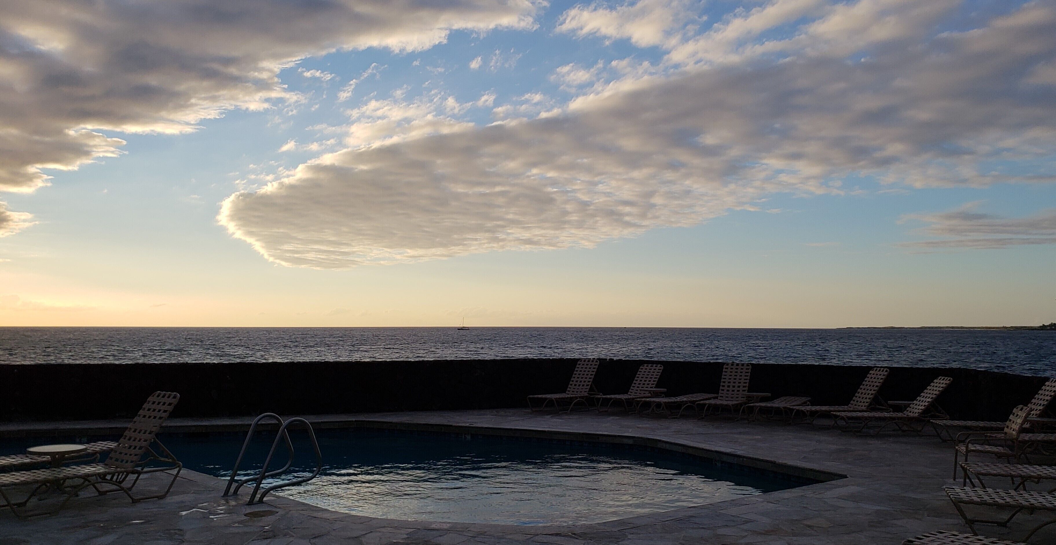 A pool area with lounge chairs and a view of the sea at sunset.