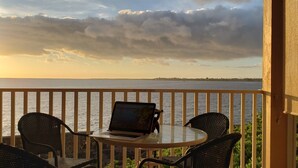 A balcony with a laptop on a table, overlooking a body of water and a distant shoreline. - Amazing Direct Ocean Front Condo - Beautiful Sunsets, watch dolphins and whales (Kailua-Kona)