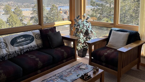 A cozy living room with a large window offering a scenic view of mountains and a lake. There is a sofa, a chair, and a coffee table with decorative items. - Views for Days Mountain Retreat.  License#6025  (Estes Park)