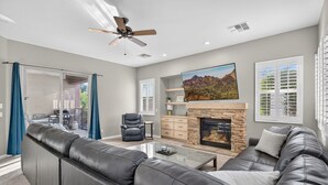 A modern living room with a fireplace, leather sofas, a flat-screen TV, and a ceiling fan.