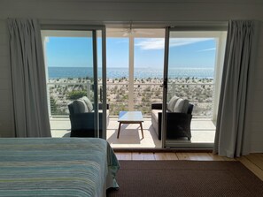 A room with a bed, a small table, two chairs, and a view of the beach and ocean through large sliding doors.