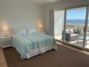 A bedroom with a bed, two bedside lamps, a view of the beach, and a balcony with a wicker chair. - Ocean Front, Sunrise Views, Well Appointed, Delaware Beach, Rehoboth By The Sea  (Dewey Beach)