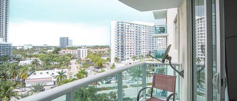 A balcony with a chair, a railing, and a view of a cityscape with tall buildings and palm trees.
