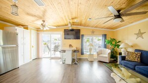 A spacious living area with a wooden ceiling, a ceiling fan, a TV, a glass coffee table, a sofa, a chair, and a kitchen area with a refrigerator and bar stools.