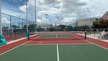 A tennis court with a net, surrounded by a fence, and a building with a thatched roof in the background.