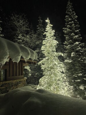 A snow-covered landscape with a prominent snow-covered tree and a cabin with a stone foundation. - Available 12/19-12/26. Luxury Home Huge Bedrooms Perfect for Multiple Families. (Park City)