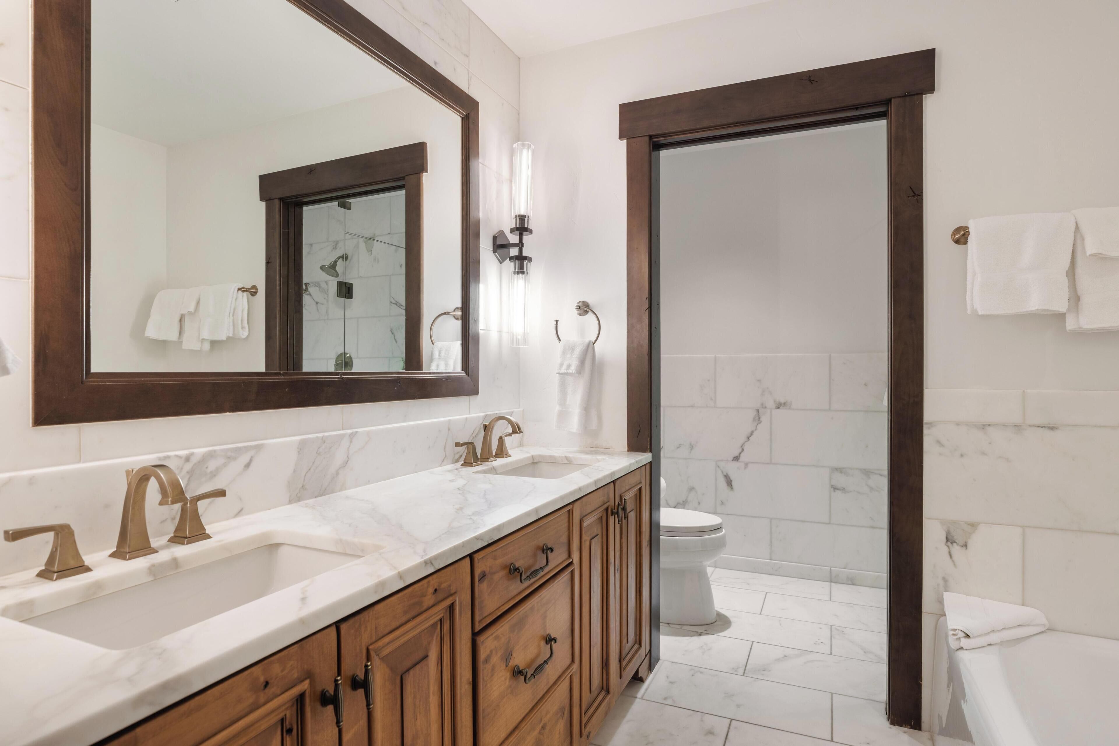 A bathroom with a large mirror, two sinks with gold faucets, wooden cabinets, and a toilet.