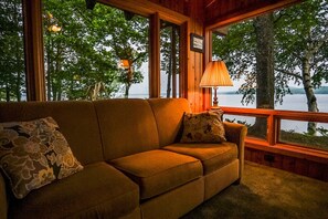 Une pièce chaleureuse avec un canapé et une lampe, de grandes fenêtres offrant une vue sur les arbres et l’eau.