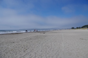 Ein weiter Sandstrand mit sanften Wellen, einem klaren Himmel und fernen Bergen.