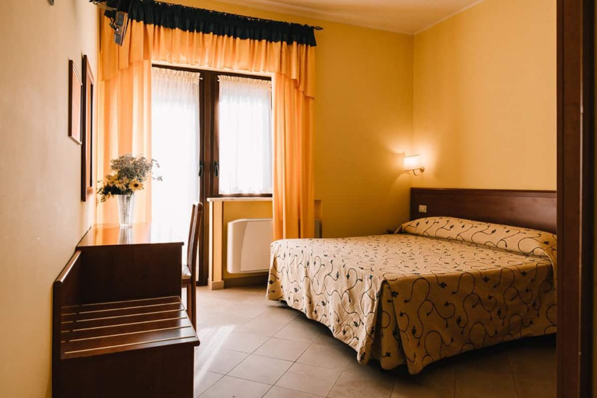 Habitación de hotel con cama, escritorio, silla, un jarrón con flores y ventana con cortinas.