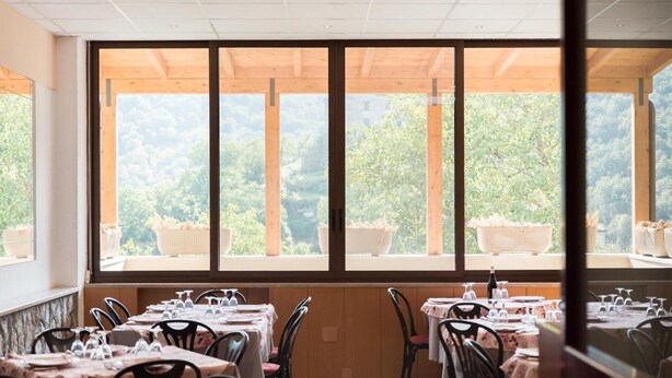 Un restaurante con grandes ventanales que ofrece vistas a un área boscosa. Las mesas están dispuestas con manteles blancos y sillas.