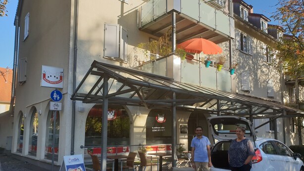 A building with a covered outdoor seating area, a car parked in front, and a sign indicating a restaurant. - Lakeside Smoker Apartment FDH (Friedrichshafen)