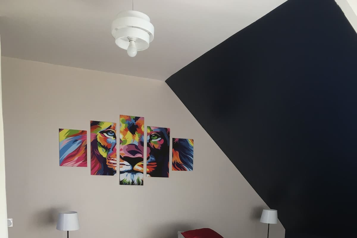 Ein Schlafzimmer mit einem Bett, einem Nachttisch, einer Lampe, einem roten Kopfkissen und einem Gemälde an der Wand.