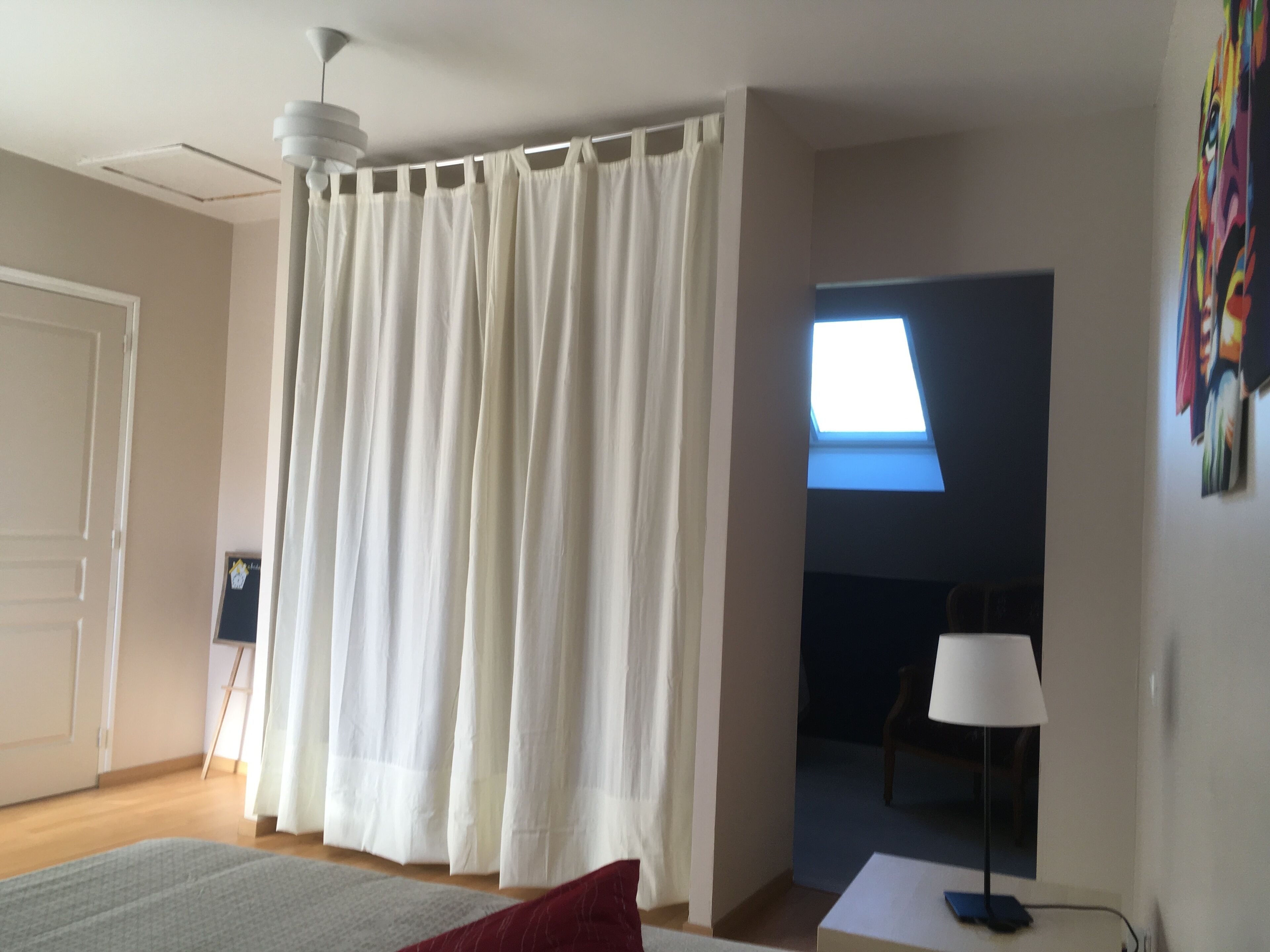 A bedroom with a bed, a white curtain, a ceiling fan, a window, and a painting on the wall.