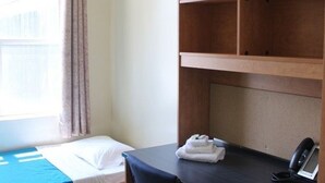 A compact bedroom with a bed, a desk, and a chair. There is a window with curtains, a bedside table with a phone, and a small storage drawer.