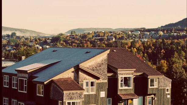 A multi-story building with a sloped roof, multiple windows, and a view of a residential area with houses and trees in the background.