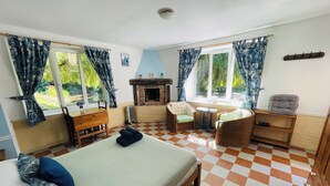A room with a bed, two chairs, a small table, a fireplace, and large windows with curtains. - Le Gite D'Ile (1 Bedroom, Sleeps 2) (Souleuvre En Bocage)