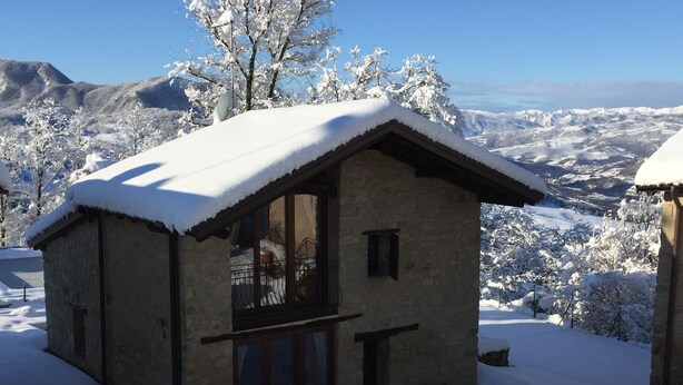 A stone building with a snow-covered roof and large windows, surrounded by snow-covered trees and a mountainous landscape. - Olimpia - Spacious stone villa with private terrace overlooking the pool (Bologna)