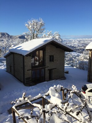 Un bâtiment en pierre, avec un toit enneigé et de grandes fenêtres, entouré d’arbres couverts de neige et d’un paysage montagneux.