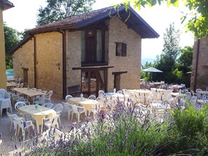 Een buitenzitplaats met stenen muren en tafels, omringd door witte plastic stoelen en paarse bloemen.