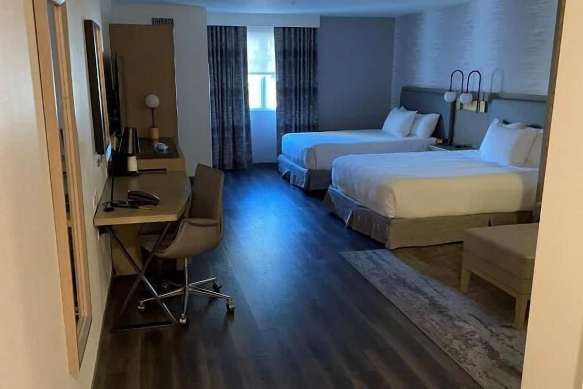 Quarto de hotel com escrivaninha, duas camas, uma cadeira e janela com cortinas.