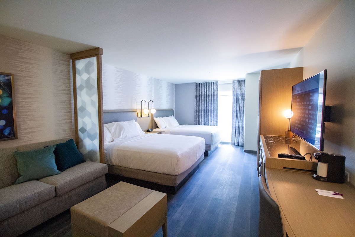 Quarto de hotel com sofá, duas camas de solteiro, escrivaninha com televisão e um espelho grande.