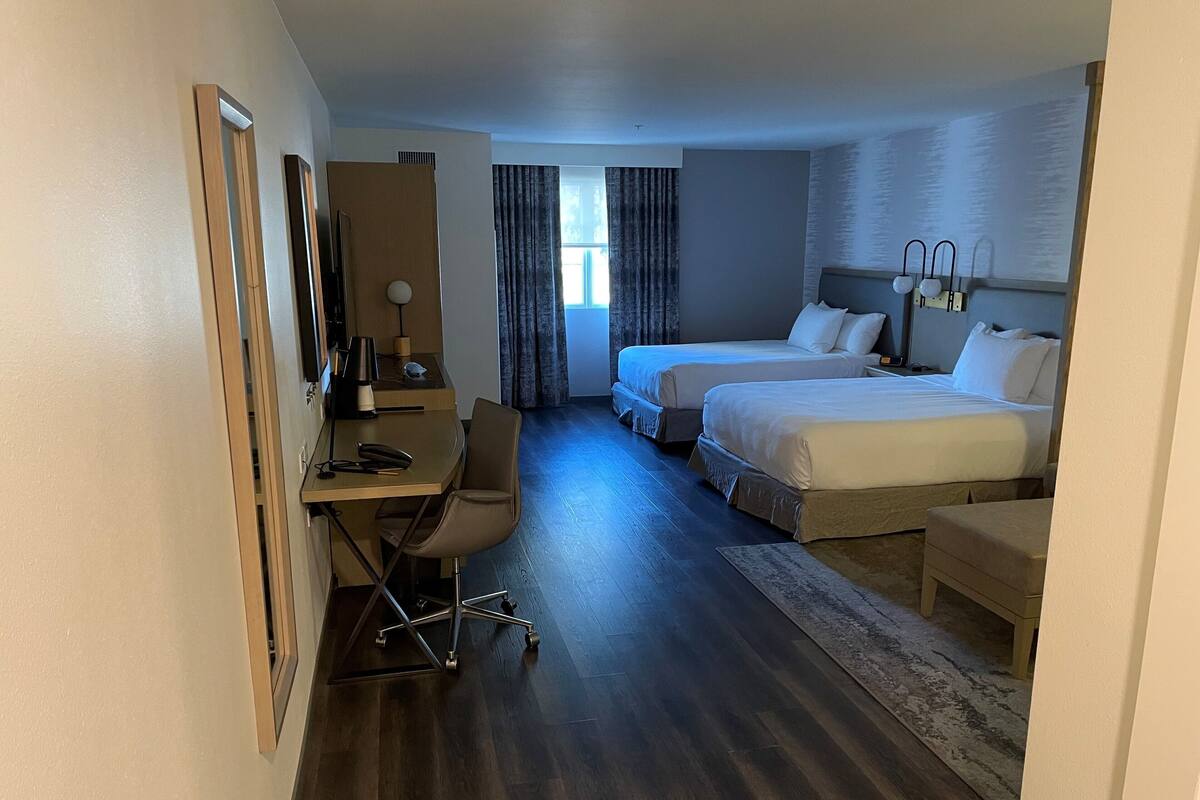 Quarto de hotel com duas camas, uma escrivaninha com cadeira, televisão e janela com cortinas.